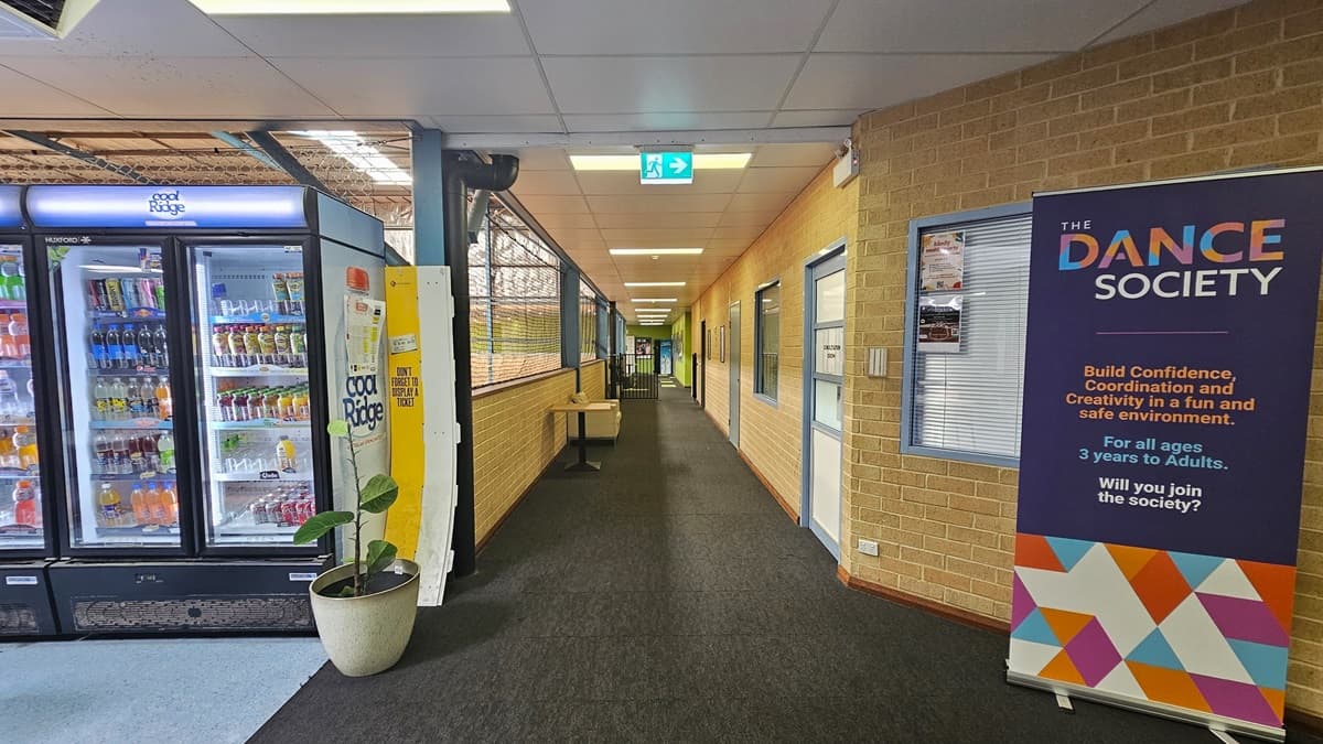 A corridor leading to other courts. On the left, a fridge for cold drinks is placed beside an ornamental plant. A table is positioned in the corridor. To the right, a large banner is near the wall. On the right, there is a brick wall with doors and windows. Further on is the entrance to the stadium, with a visible exit sign and other banner ads. The floor has grey carpets in the middle and blue vinyl on the left.