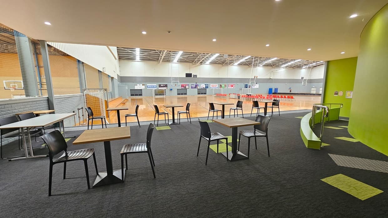 In the middle, plastic chairs and tables are arranged. To the right, a ramp with handrails and truncated domes is present. Behind is the court with timber floors and an industrial ceiling with exposed beams. The floor is covered in grey carpet with a neon green accent. The right wall is painted neon green.