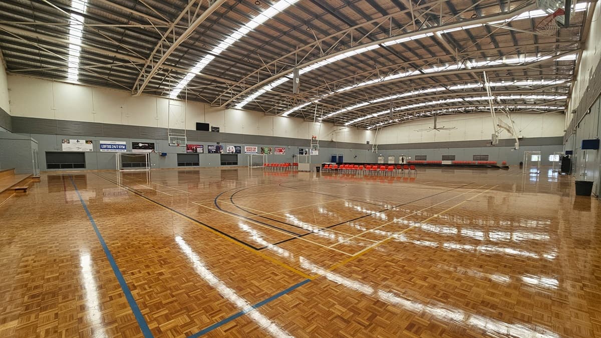 The stadium features timber floors marked for various ball games. Plastic red chairs are arranged in the middle, separating the two courts. Large windows line the walls, accompanied by banners and a digital screen. The industrial ceiling has exposed metal beams and reflective insulation. Fluorescent lights contribute to glare and reflections on the timber floor.