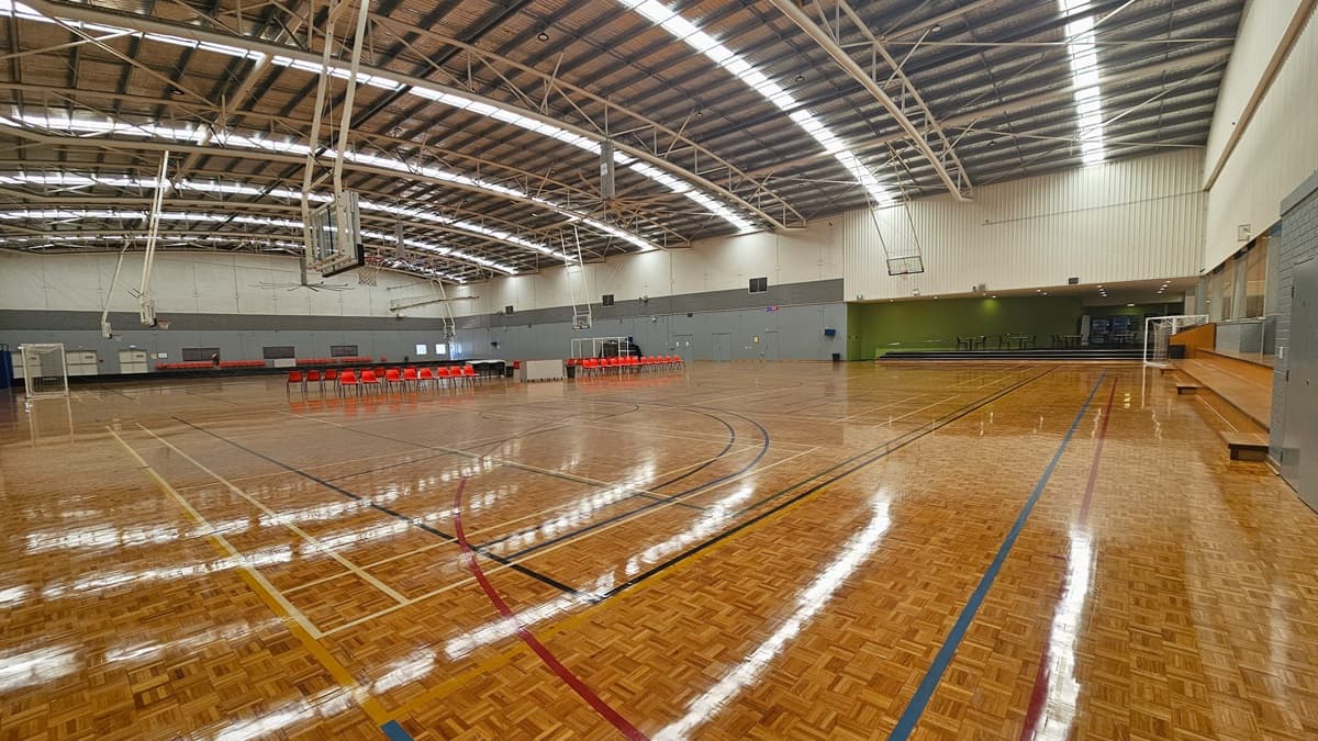 The stadium has timber floors marked for various ball games. Plastic red chairs are arranged in the middle, dividing the two courts. Large windows line the walls, with the upper wall in white and corrugated design. The industrial ceiling features exposed metal beams and reflective insulation. Fluorescent lights contribute to glare and reflections on the timber floor. Further right, a step leads to the area with arranged plastic tables and chairs.