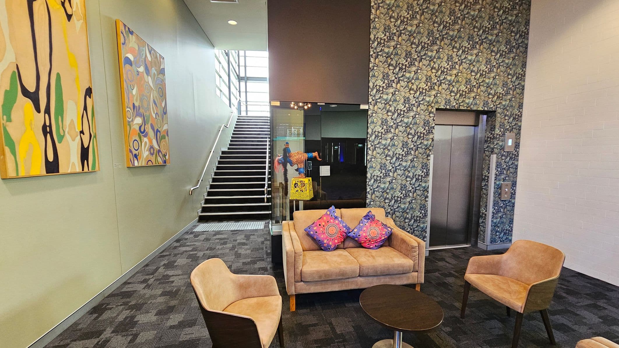 A modern interior space with a staircase on the left, featuring two abstract paintings on the wall. In the centre, a glass partition displays a colourful artwork. A seating area includes a beige sofa with two patterned cushions and two matching chairs around a small round table. On the right, an elevator is set into a wall with a floral pattern. The floor is carpeted with a dark geometric design.