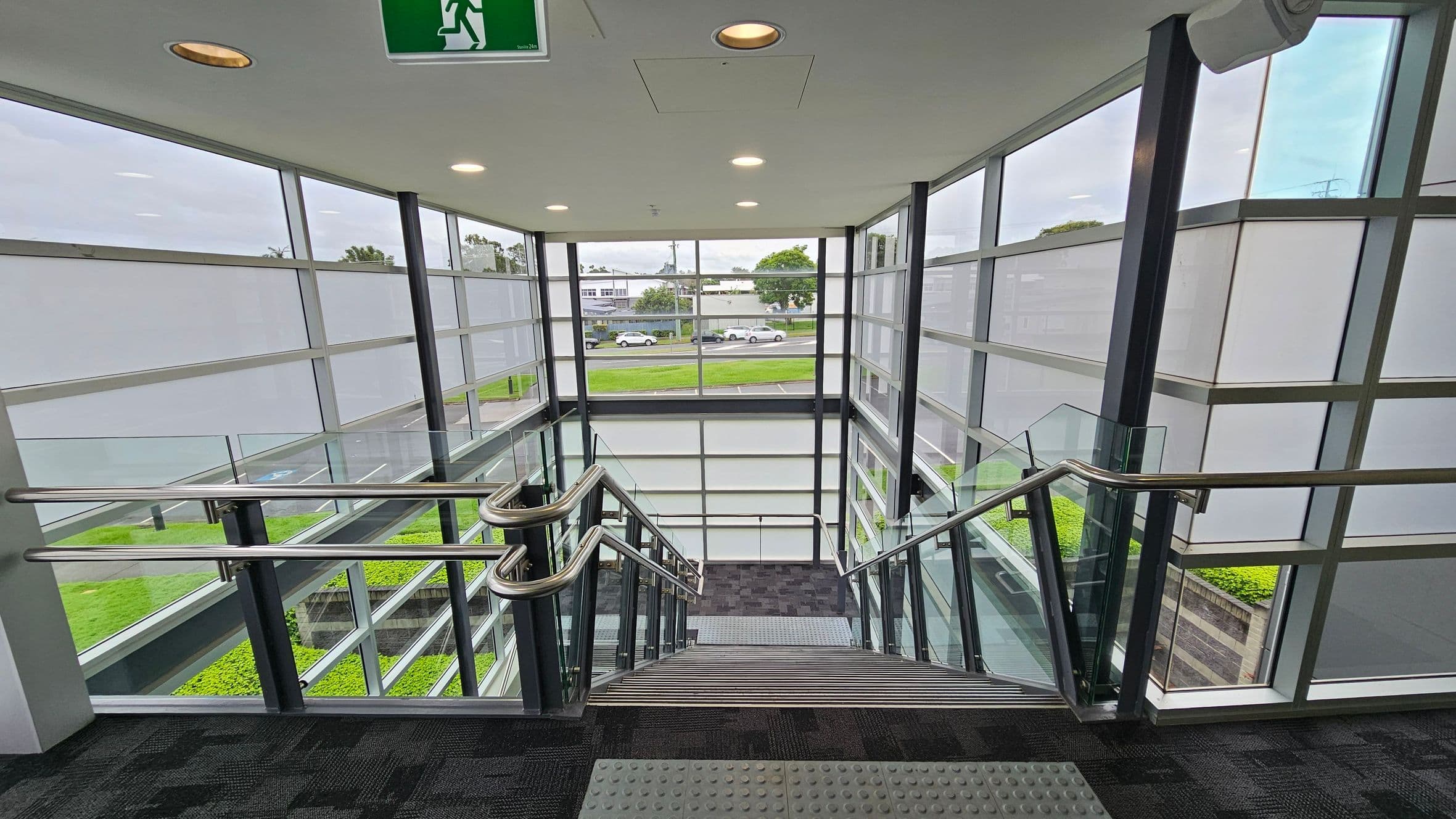 A modern indoor staircase with metal railings and glass panels on both sides. The walls are made of large glass windows, providing a view of the outside, including a road with parked cars and greenery. The ceiling is white with recessed lighting and an emergency exit sign. The floor is carpeted in a dark pattern, and there are tactile paving tiles at the top of the stairs.