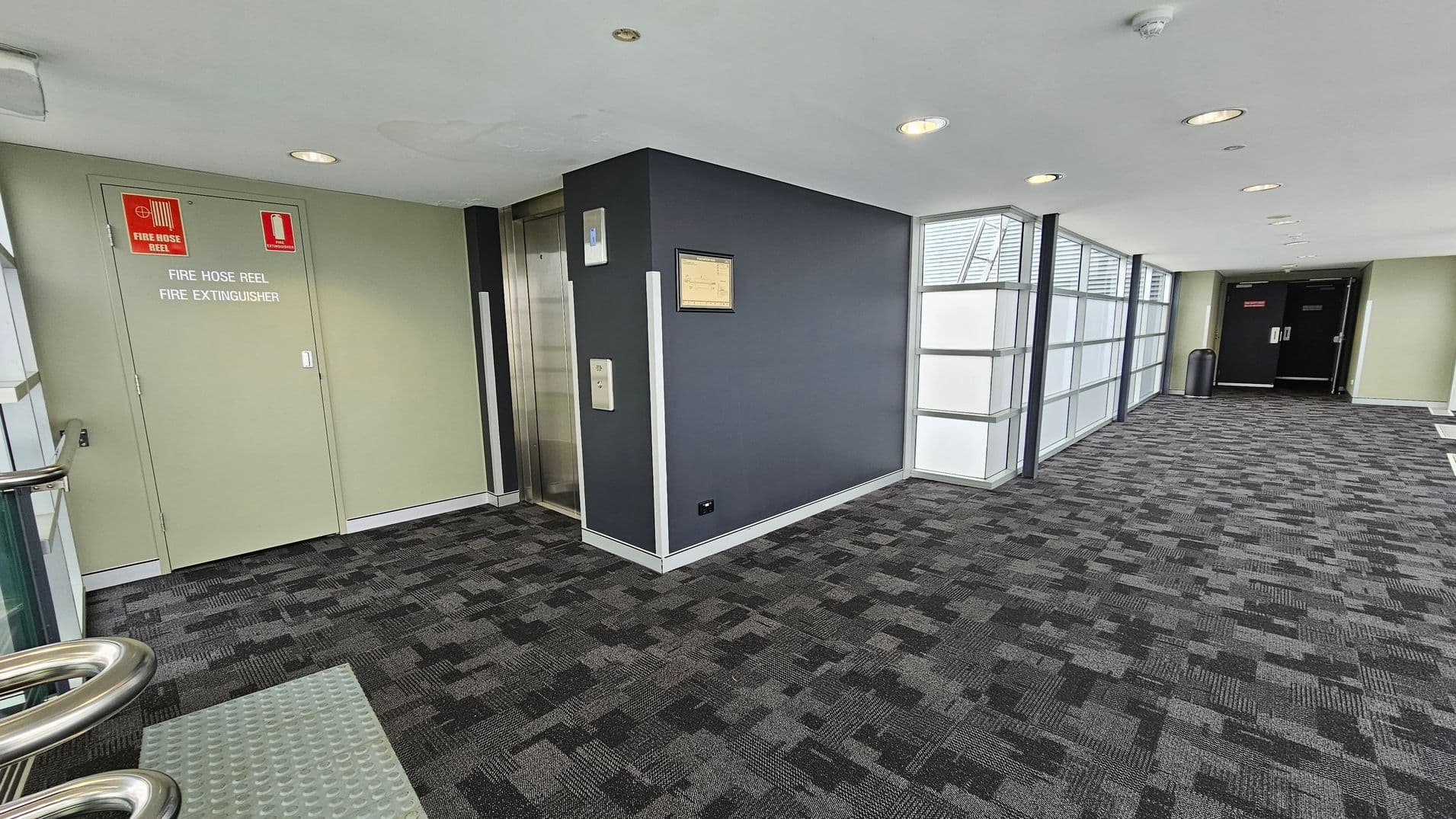 A corridor with a patterned grey carpet. On the left, there is a green door marked for a fire hose reel and extinguisher. Next to it is a lift with a metallic door. The wall beside the lift is dark grey with a framed notice. On the right, large windows with frosted glass panels allow light in. At the end of the corridor, there are double black doors with a bin nearby. The ceiling is white with recessed lights.