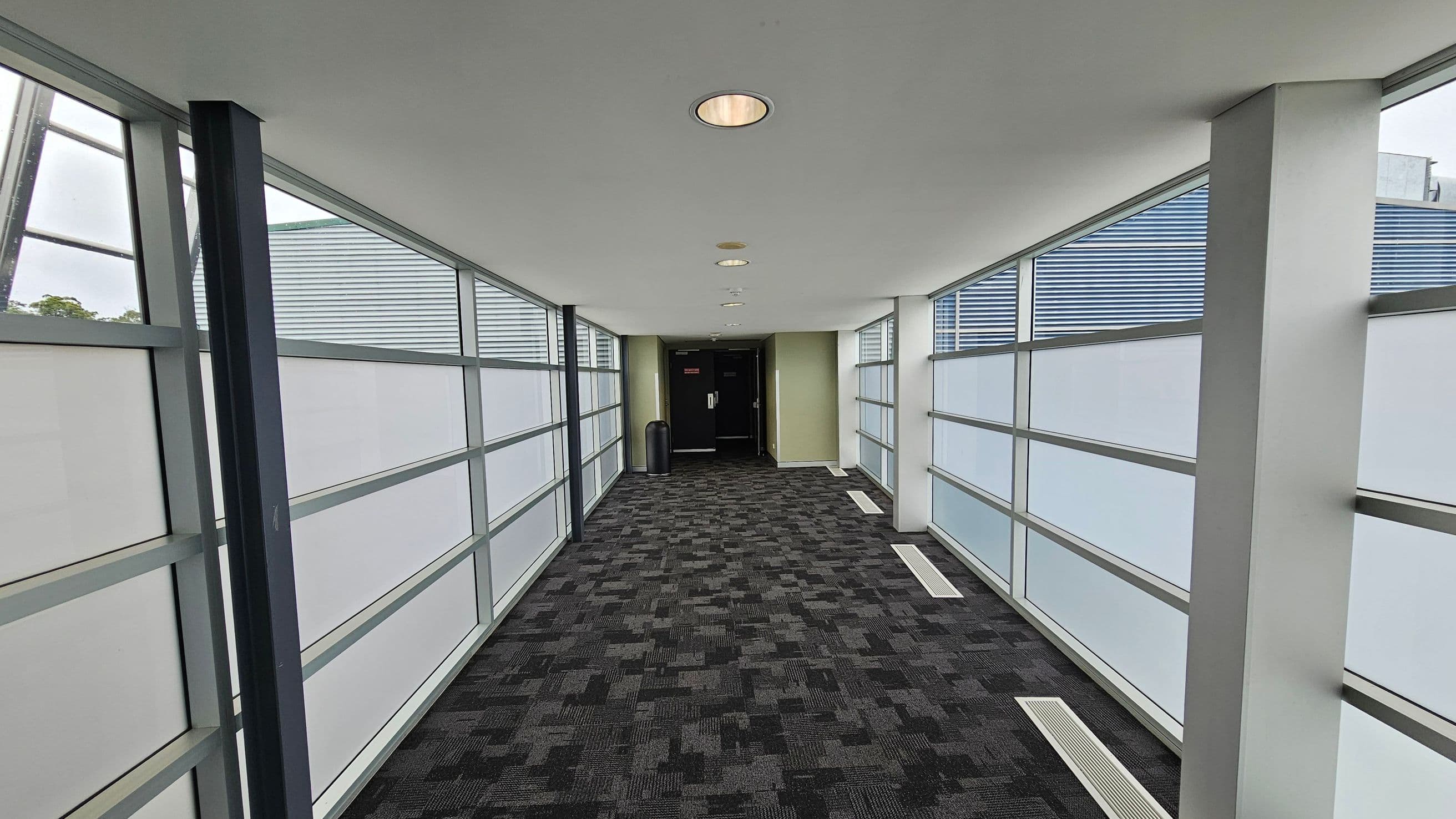 A corridor with large frosted glass windows on both sides, framed by metal supports. The floor is covered with a dark patterned carpet. The ceiling is white with recessed circular lights. At the end of the corridor, there is a black door and a green wall, with a black bin nearby.