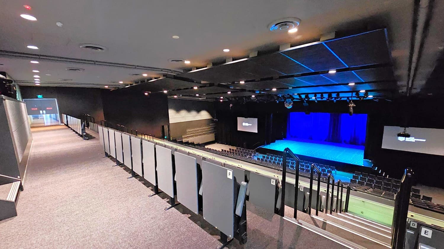 A theatre interior with a view from the upper seating area. On the left, a carpeted walkway leads to an exit door. The right side features tiered seating with grey panels and handrails. In the middle, a stage with a blue-lit backdrop is visible, flanked by two large screens. The ceiling is equipped with recessed lighting and ventilation ducts. The floor is carpeted, and the overall atmosphere is dimly lit.