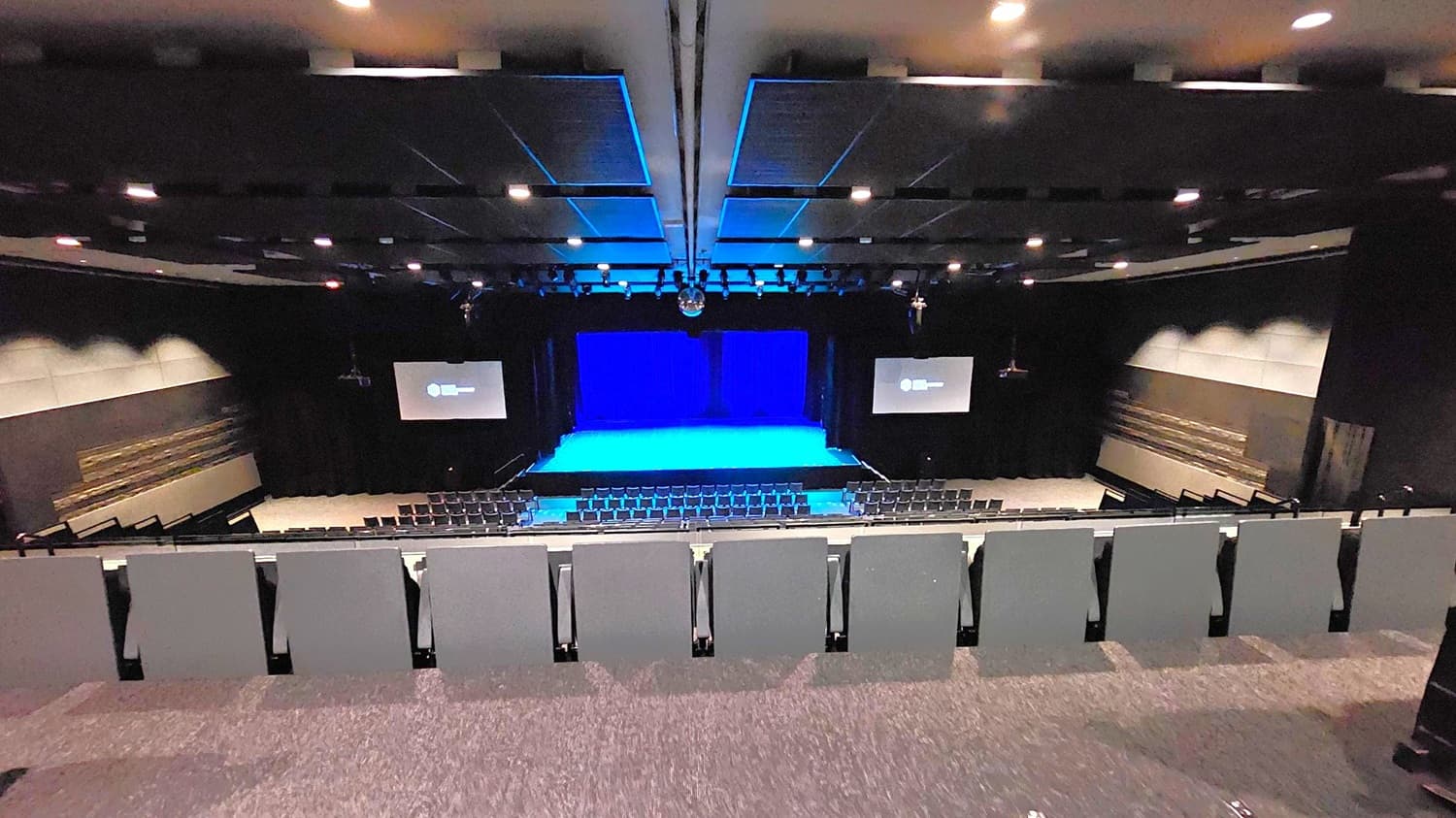 A large auditorium with tiered seating in the foreground, leading down to a stage with a blue-lit backdrop. Two screens are positioned on either side of the stage, displaying a logo. The walls are dark with acoustic panels, and the ceiling is equipped with numerous recessed lights. The floor is carpeted in a dark colour, matching the overall dim and professional atmosphere of the space.