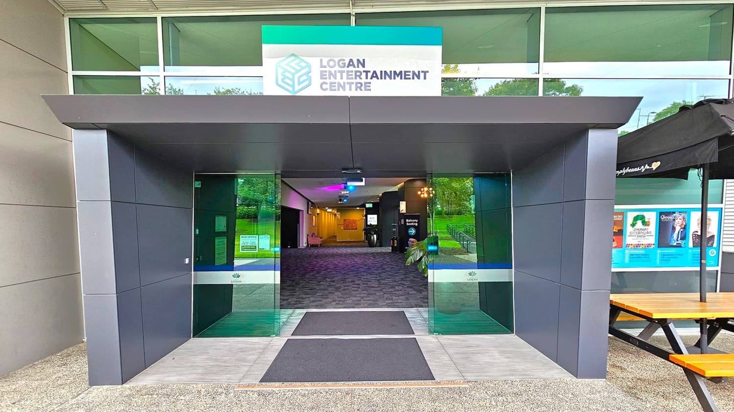 A modern entrance to the Logan Entertainment Centre with a grey and green colour scheme. The entrance features large glass doors with a sign above displaying the centre's name and logo. Inside, a carpeted hallway is visible, leading to various rooms. To the right, there is a wooden picnic table and a display of posters under a black canopy. The exterior walls are clad in grey panels, and the ground is covered with a textured surface.
