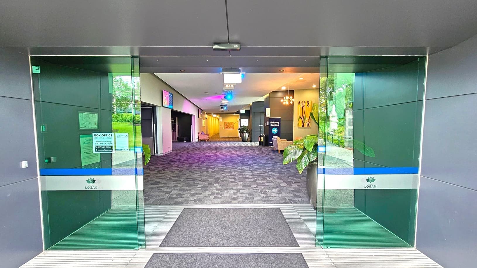 Glass doors with a blue and white stripe, labelled "Logan," open into a spacious lobby. The floor is covered with a patterned carpet. On the right, a potted plant and a sign for "Balcony Seating" are visible. The left side has a box office window with opening hours displayed. The ceiling features recessed lighting and colourful spotlights. In the background, there are more seating areas and artwork on the walls.