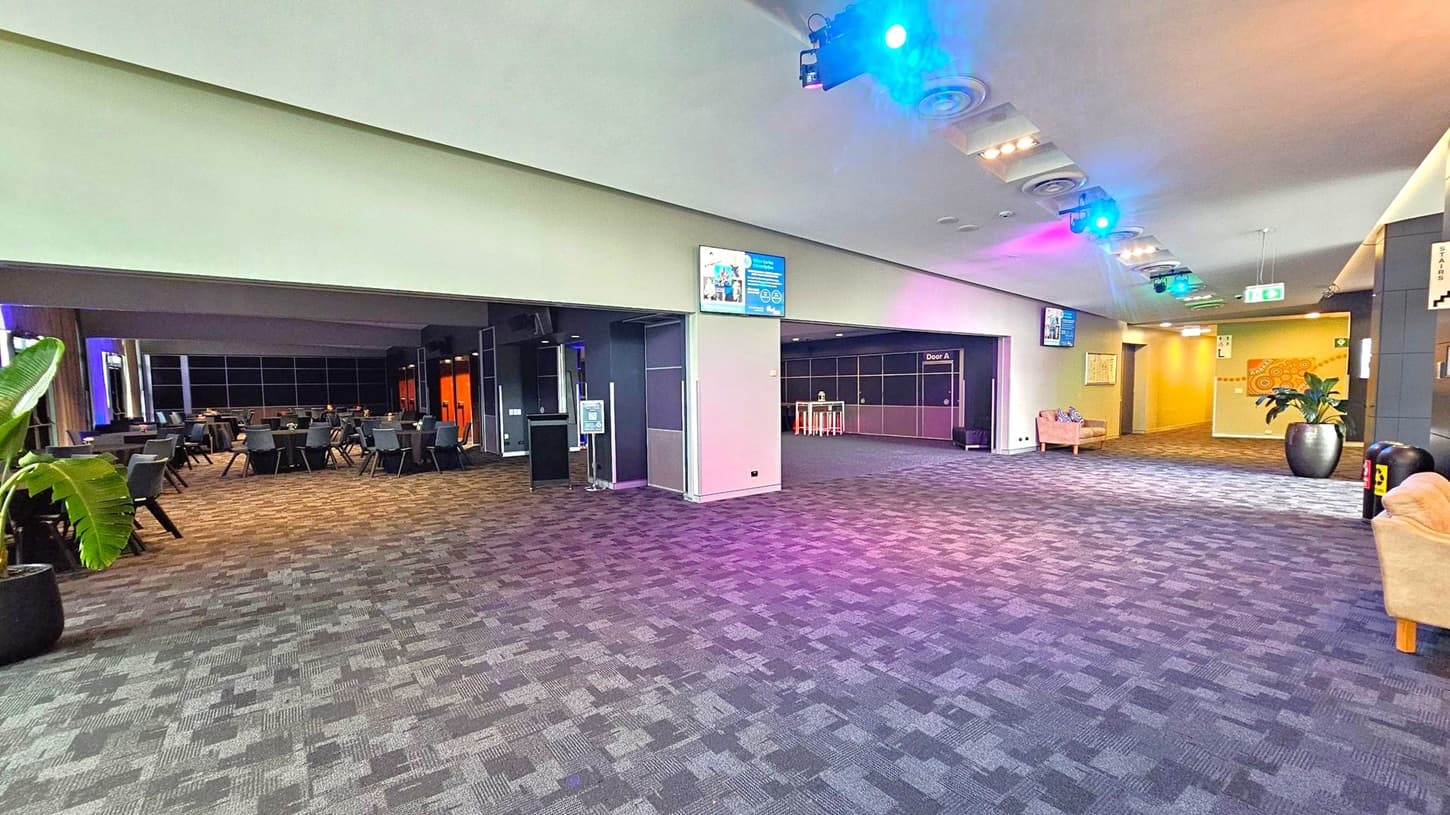 A spacious indoor area with a patterned carpet floor. On the left, there are tables and chairs arranged in a dining setup, with a large potted plant nearby. The right side features a hallway with a large potted plant and a beige sofa. The ceiling has recessed lighting and coloured spotlights. Digital screens and signs are mounted on the walls, and there are doors labelled "Door A". The walls are decorated with abstract art.