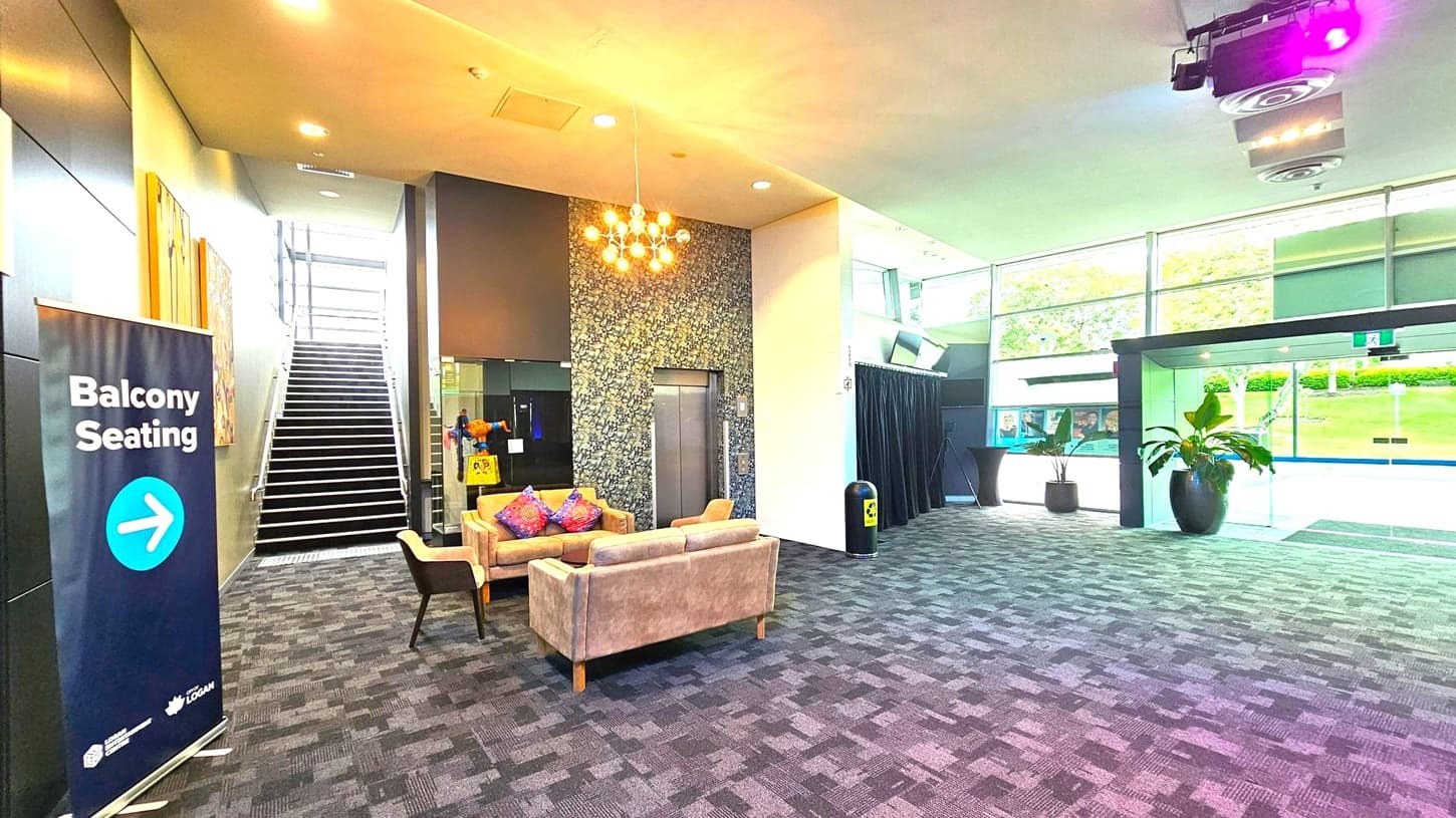 A modern interior space with a patterned carpet floor. On the left, a sign indicates "Balcony Seating" with an arrow. A staircase with metal railings leads upwards. In the centre, a seating area features a beige sofa and chairs with colourful cushions, under a cluster of hanging lights. An elevator is nearby. On the right, large windows provide a view of greenery outside. A potted plant and a small table are near the glass entrance doors. The ceiling has recessed lighting and a purple spotlight.