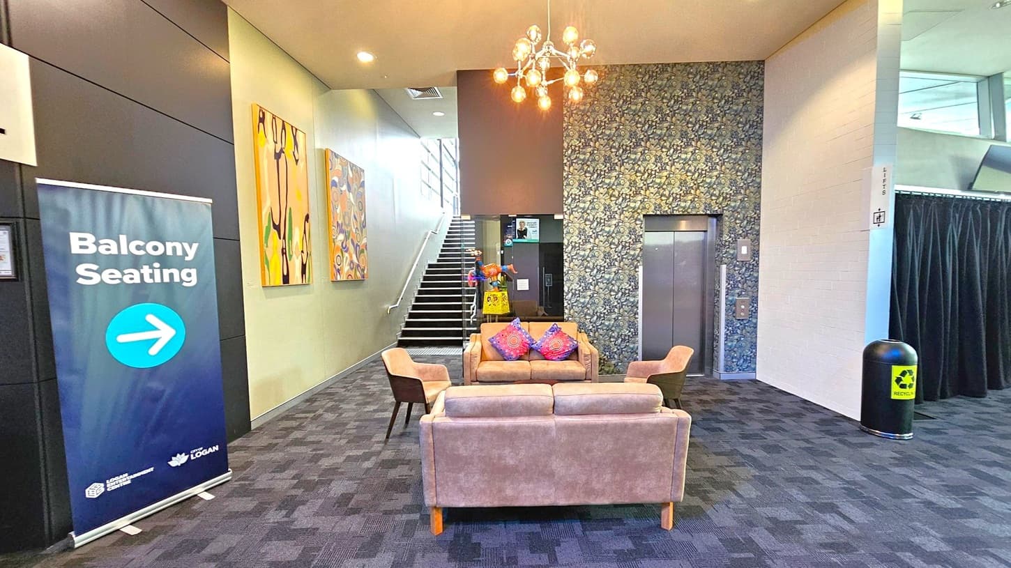 A modern interior space with a patterned carpeted floor. In the centre, there are two beige sofas and two chairs with colourful cushions. A chandelier hangs from the ceiling. On the left, a sign reads "Balcony Seating" with an arrow. Behind the seating area, a staircase leads upwards, flanked by abstract art on the wall. To the right, there's a lift with a floral-patterned wall and a recycling bin nearby.