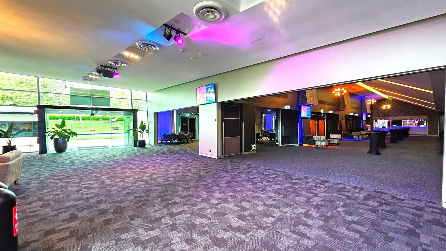 Spacious interior with a patterned carpet floor. On the left, large glass doors lead outside, flanked by potted plants. The ceiling features circular vents and colourful lights. On the right, an open area with tables and chairs, illuminated by warm lighting. A bar is visible in the background. Monitors are mounted on the walls, and a sofa is partially visible on the far left.