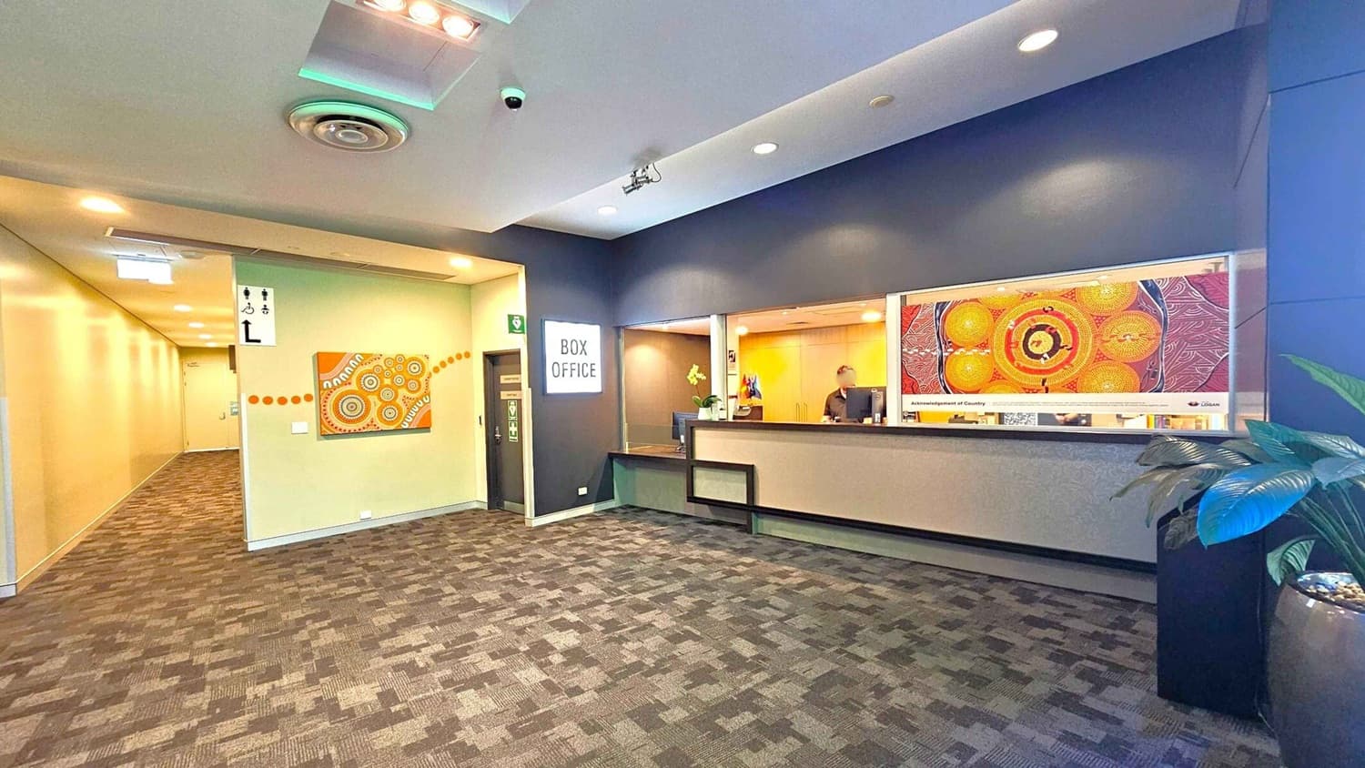 A modern interior with a box office on the right, featuring a counter and a person behind it. Above the counter is a colourful artwork. On the left, a hallway extends with a patterned carpet and a wall adorned with another piece of art. The ceiling has recessed lighting and a circular vent. A potted plant is in the foreground on the right. The floor is covered with a patterned carpet.