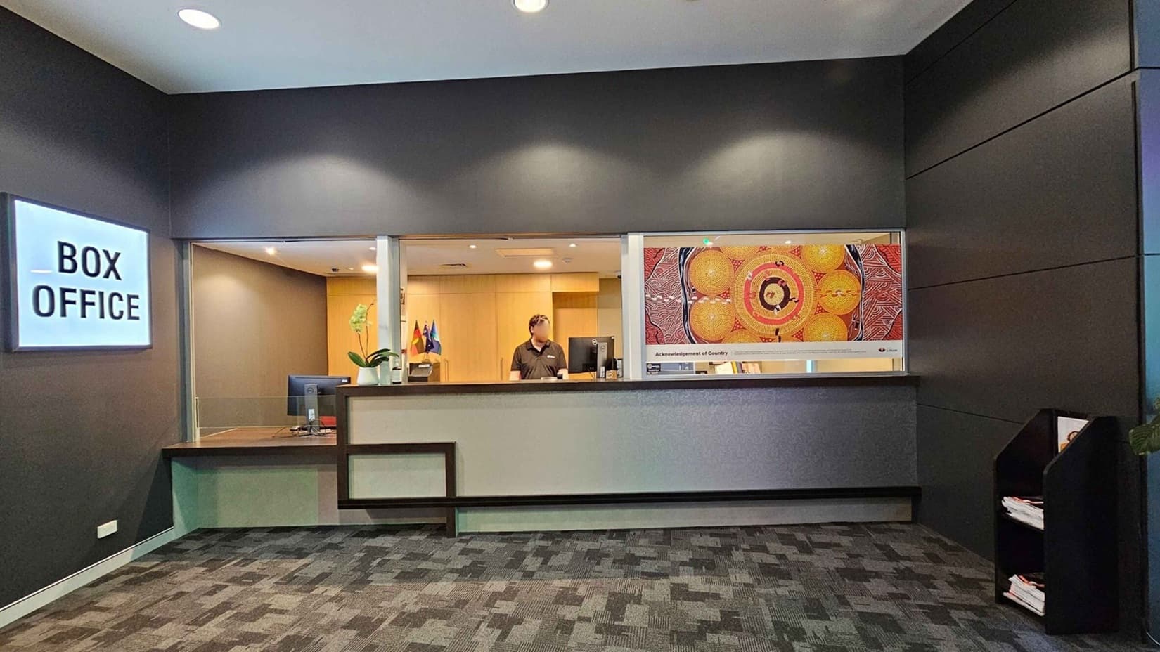 A box office area with a grey and black colour scheme. On the left, a lit sign reads "BOX OFFICE" above a counter with a computer and a potted plant. In the centre, a person stands behind the counter, with flags visible in the background. On the right, a colourful artwork is displayed on the wall. Below the artwork, a shelf holds brochures or papers. The floor is carpeted with a patterned design, and the ceiling has recessed lighting.