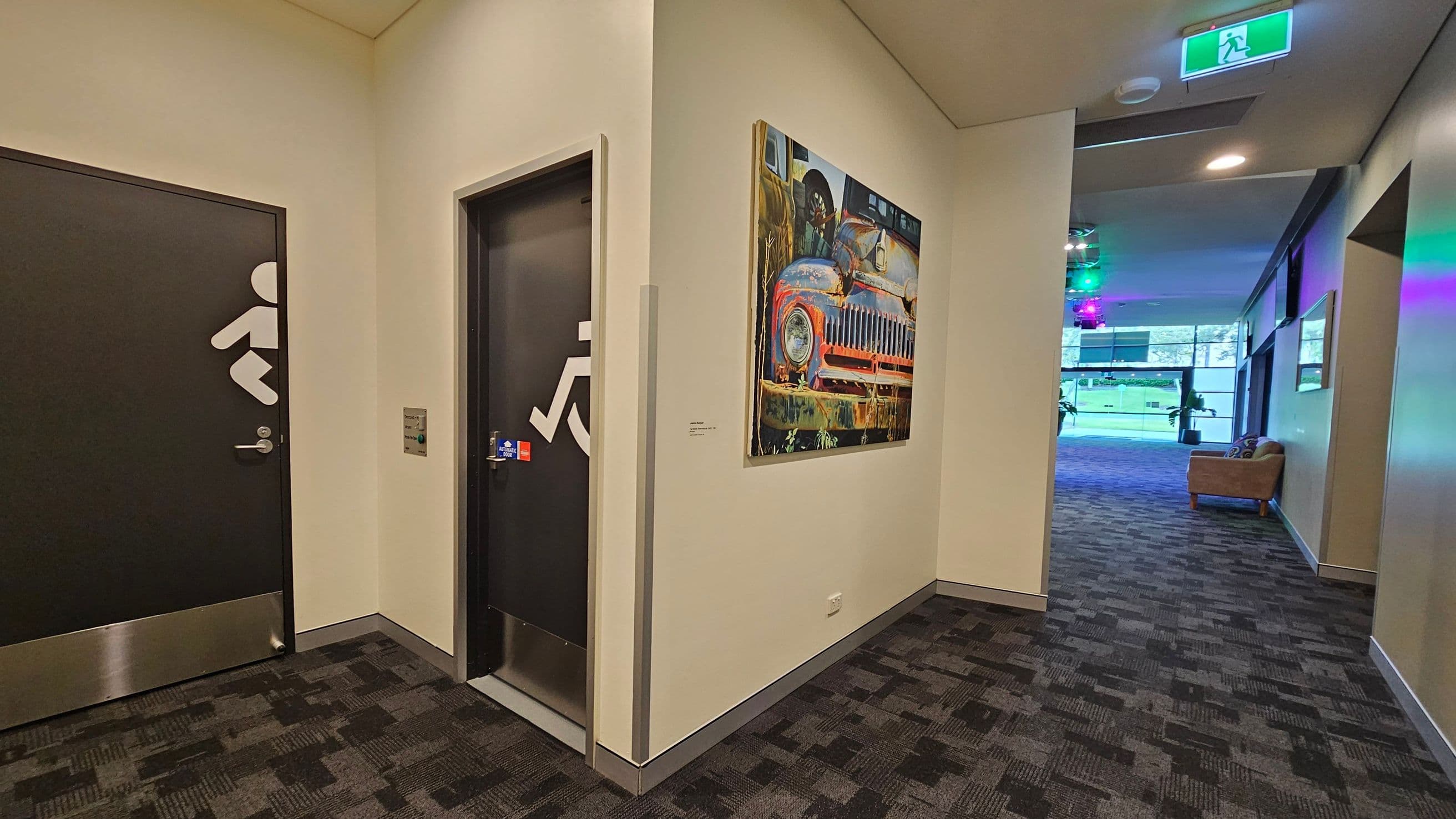 A hallway with two black doors on the left, each marked with white symbols indicating accessibility. Between the doors, a colourful painting of an old car is mounted on the wall. The floor is covered with a patterned carpet. To the right, the hallway opens into a larger room with a sofa and large windows. The ceiling has recessed lighting and an illuminated green exit sign.