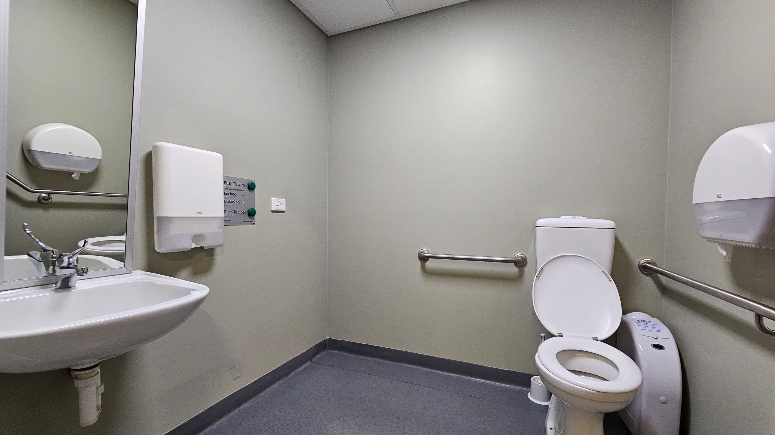 A public restroom with grey walls and a light grey ceiling. On the left, a white sink with a metal tap is mounted below a large mirror. A paper towel dispenser is above the sink. In the centre, a wall-mounted panel with buttons for locking and unlocking. On the right, a white toilet with the lid open, flanked by metal grab bars. A toilet paper dispenser is mounted on the wall. The floor is covered with dark grey tiles.