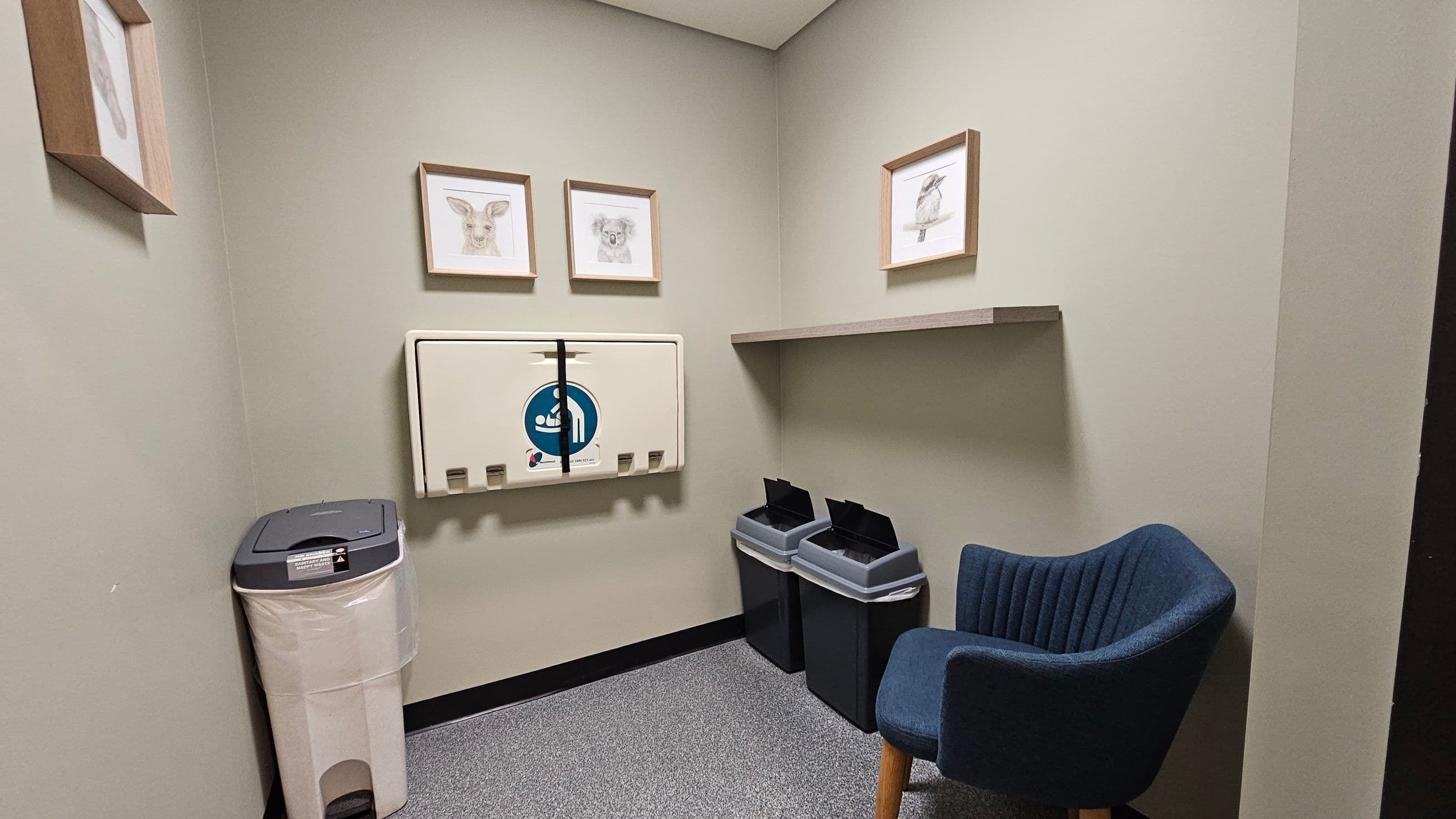 A small room with light green walls. On the left, a beige nappy changing station is mounted on the wall, with a waste bin below it. Above the station are three framed animal sketches. On the right, a wooden shelf is mounted on the wall, with two black bins beneath it. A blue upholstered chair with wooden legs is positioned in the corner. The floor is covered with grey speckled carpet tiles.