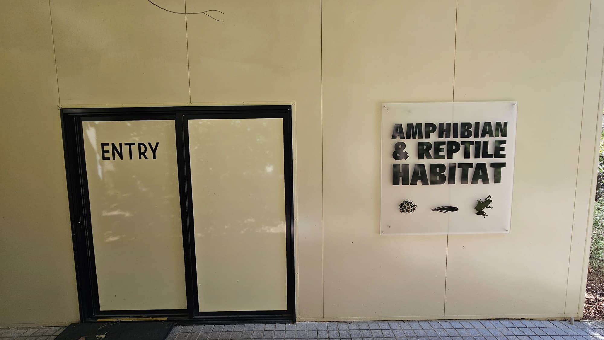 Entrance of the amphibian and reptile habitat. A sign with “Amphibian & Reptile Habitat” with images of the life cycle of a frog is prominent on the right. Next to it is a sign that depicts an entrance in a black hue with “ENTRY” in it. The floor surface clearly features a brick pathway.