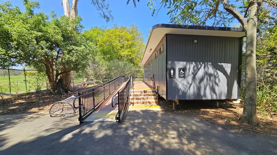 On the right, there's an accessible toilet block featuring grey corrugated walls. Stairs and a ramp with railings, both equipped with yellow truncated domes, are situated on the left. A signage indicating accessibility and gender access is mounted on the wall. Adjacent to the ramp on the left is a dome-shaped metal cage enclosed by a chain metal fence. The surroundings are dominated by large, mature trees.