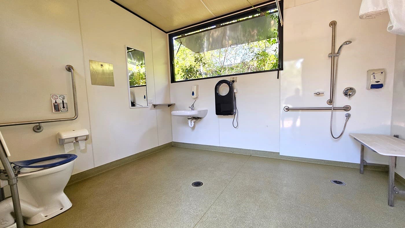 In the accessible bathroom, a toilet bowl with metal handrails and a tissue dispenser is placed in the left corner. On the left wall, there's a mirror. A large open window is on the right. A wall-mounted washbasin, liquid soap dispenser, and hand dryer are present. Adjacent is a shower with handrails and another tissue dispenser, all affixed to the wall. A white bench is positioned near the corner. The floor is tiled with granite, and the walls are white.