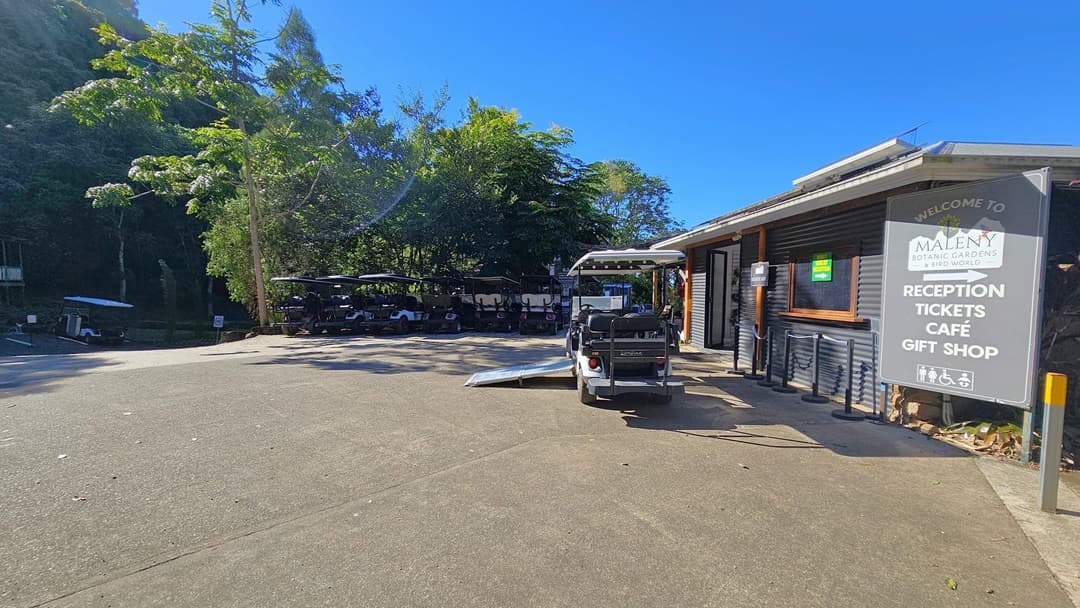On the left, there are several golf carts parked under the shade of tall trees. In the middle, there is a concrete pathway leading to a building with a wooden facade. The building has a sign listing reception, tickets, café, and gift shop. On the right, there is a golf cart parked near the entrance of the building. The floor is concrete, and the building has a grey roof with a slight overhang. The background features dense greenery and more trees.
