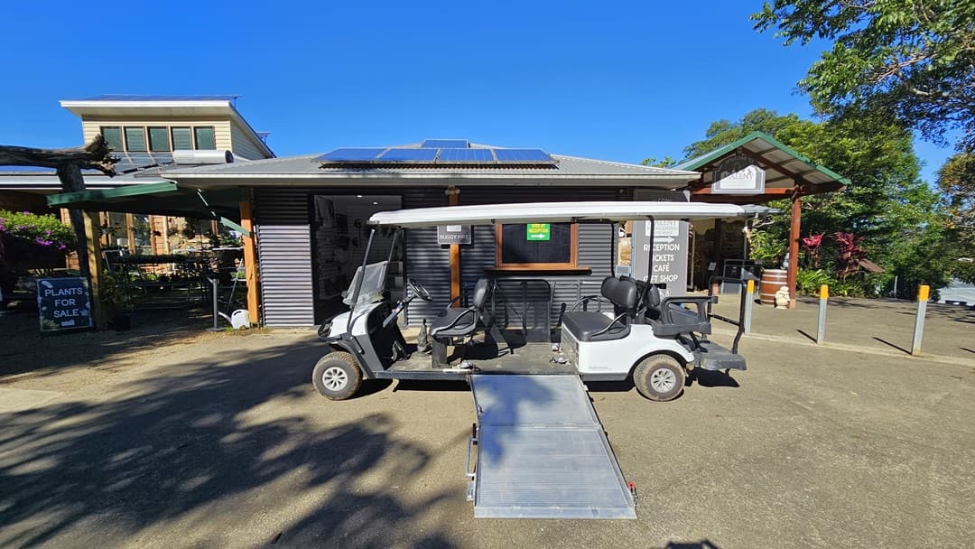 On the left, there is a sign that reads "Plants for Sale" next to a shaded area with plants. In the middle, there is a building with a grey roof featuring solar panels, a wooden facade, and siganges. A golf cart with a ramp is parked in front of the building. On the right, there is a covered walkway with a wooden post and bollards. The floor is concrete, and the ceiling of the shaded area and walkway is wooden. The background includes trees and plants.