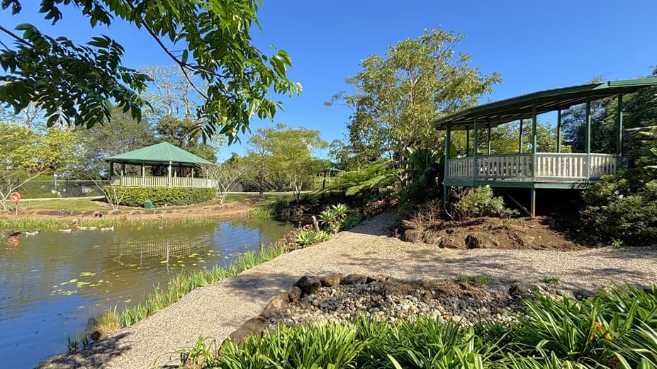 On the left, there is a pond with water lilies and a green-roofed gazebo near the water's edge. In the middle, there is a gravel path bordered by rocks and plants, leading towards the pond. On the right, there is an elevated green gazebo with a white railing, surrounded by trees and bushes. The background includes more trees and greenery. The floor is covered with gravel and grass.