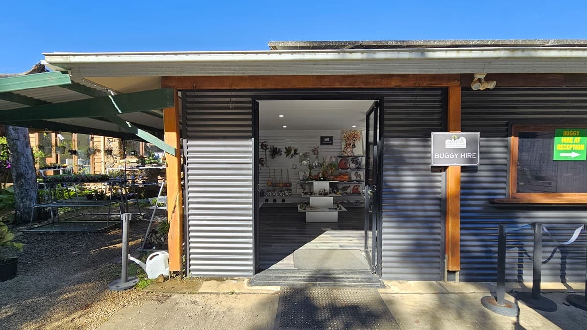 A low-set building with a corrugated front and closed window, its door open to reveal retail products on display. In the middle, a display rack holds plants for sale, accompanied by a sign. To the left, two golf buggies are parked under the shade of trees. The ground is covered with gravel, transitioning to concrete on the right. Signages are visible in the background.