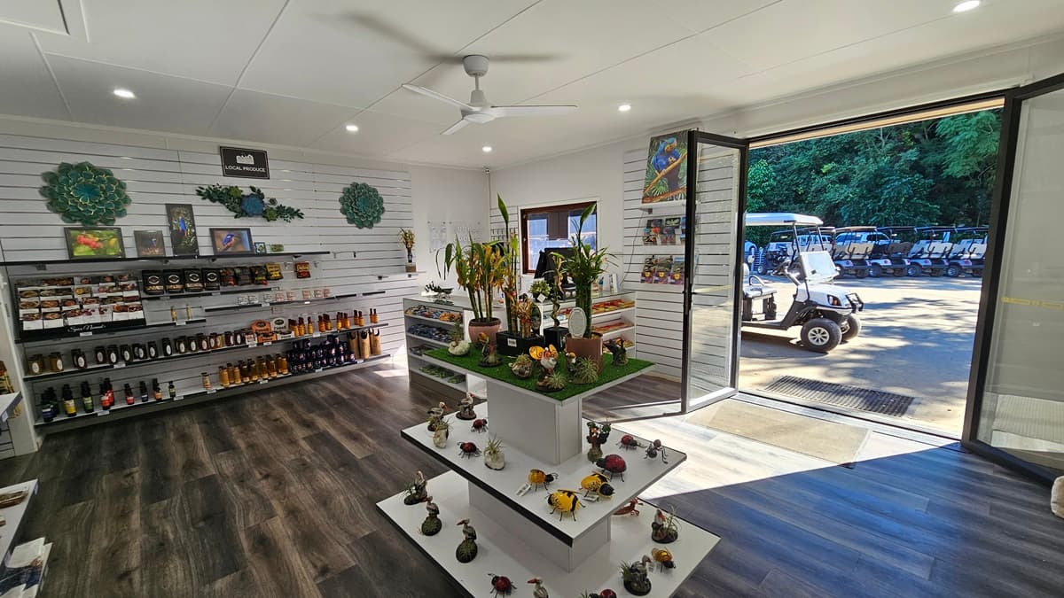 Retail items including local arts, crafts, and gourmet sauces are arranged on display racks against the walls and in the middle of a room with dark timber flooring and white brick walls. The ceiling is white, featuring a ceiling fan and recessed lighting. To the right, a door opens inward, equipped with a non-slip mat. Outside, golf buggies and dense greenery are visible.