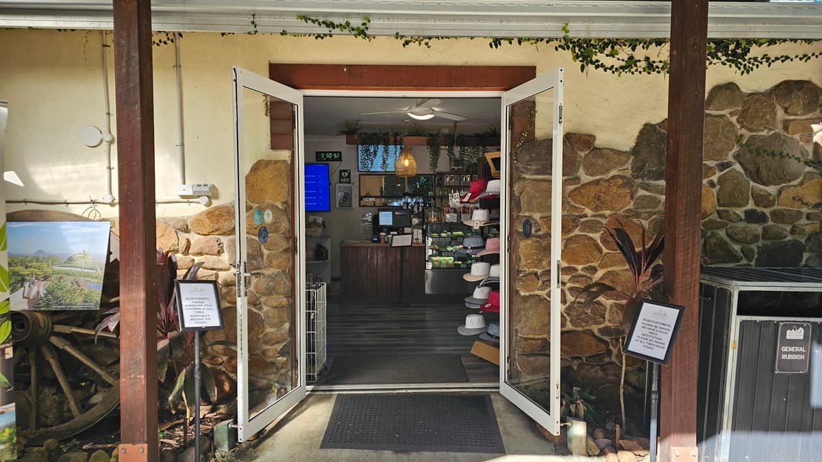 An outward-opening glass door reveals a section of a room displaying retail items such as hats, caps, and artworks. To the left, a large wooden wheel decoration holds a painting. On the right, a segregation rubbish bin and signages are present. The building's outer wall features a cobblestone design with cream paint. The flooring transitions from concrete outside to timber inside.