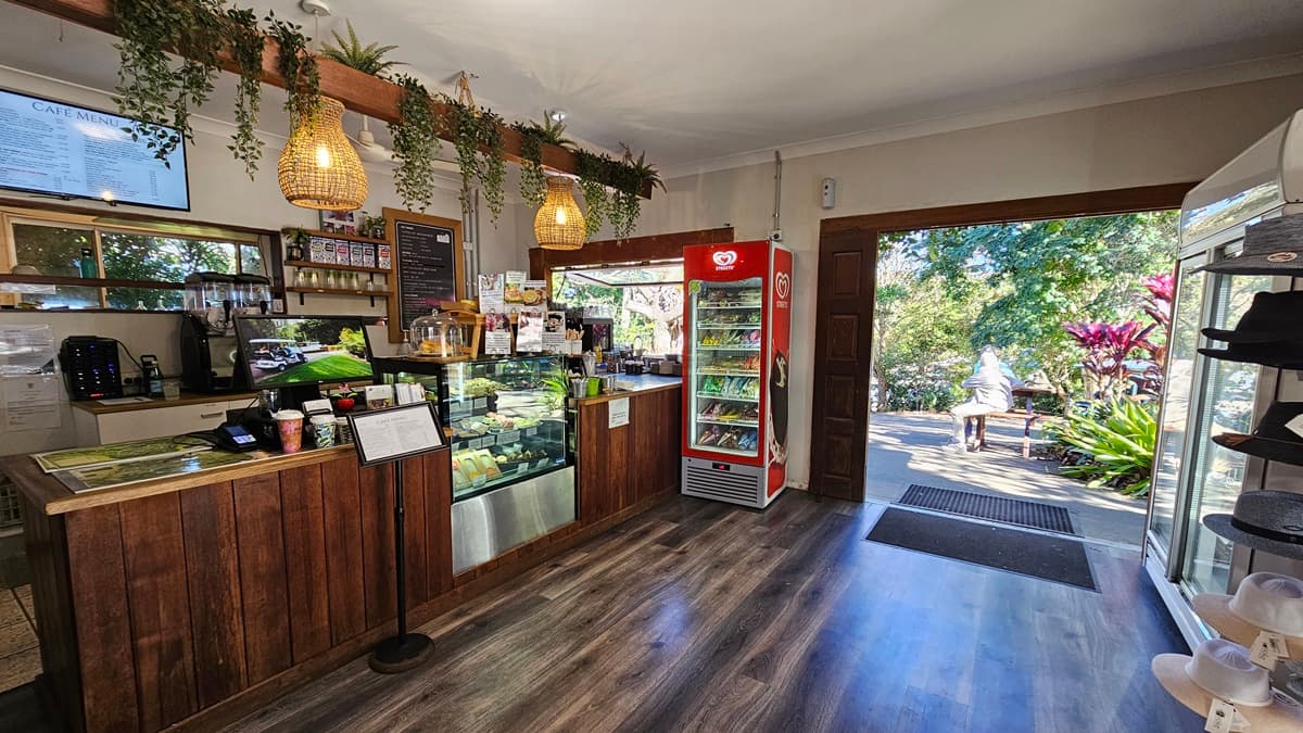 On the left, there is a wooden counter with a glass display case containing food items. Above the counter, hanging plants and pendant lights are visible. Behind the counter, there are shelves with various items and a menu board. On the right, there is a red refrigerator stocked with drinks and a white refrigerator with more items. In the middle, a doorway leads outside to a garden area with trees and plants. The floor is wooden, and the ceiling is white with recessed lighting.