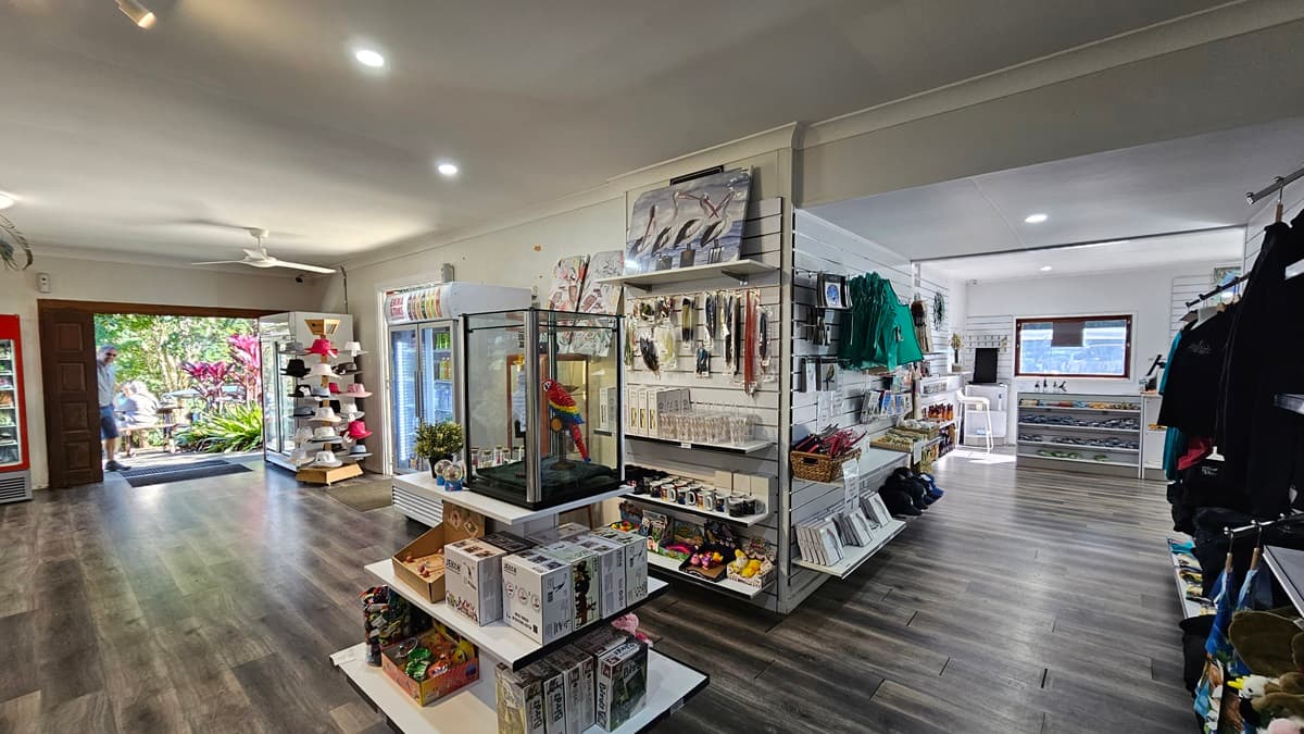 In the middle, there are shelves with various items, including books and toys, and a glass display case with a colourful parrot model. On the left, there is an open doorway leading to a garden area with plants and a seating area. On the right, there are more shelves with clothing and accessories, and a counter with additional items. The floor is dark wood, and the ceiling is white with recessed lighting and a ceiling fan.