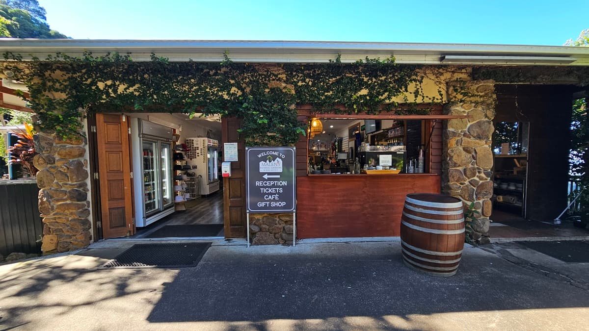 On the left, there is an open wooden door leading to an interior with shelves displaying various items. In the middle, there is a wooden counter with a sign listing reception, tickets, café, and gift shop, and a window with a view of the interior. On the right, there is a large wooden barrel and another open doorway. The background features stone walls with green vines. The floor is concrete, and the ceiling is white with a slight overhang.