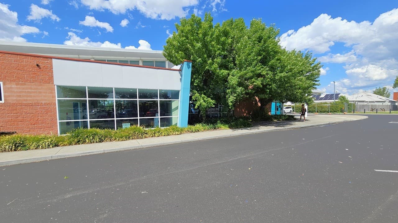 The facility building is a commercial structure featuring red brick, blue-colored concrete, steel, and glass elements. The left side of the building is accompanied by trees and ornamental plants, running parallel to the building and the concrete pathway. The entrance is further ahead, leading to additional commercial buildings equipped with solar panels. A person is standing under the shade of a tree near the curb.