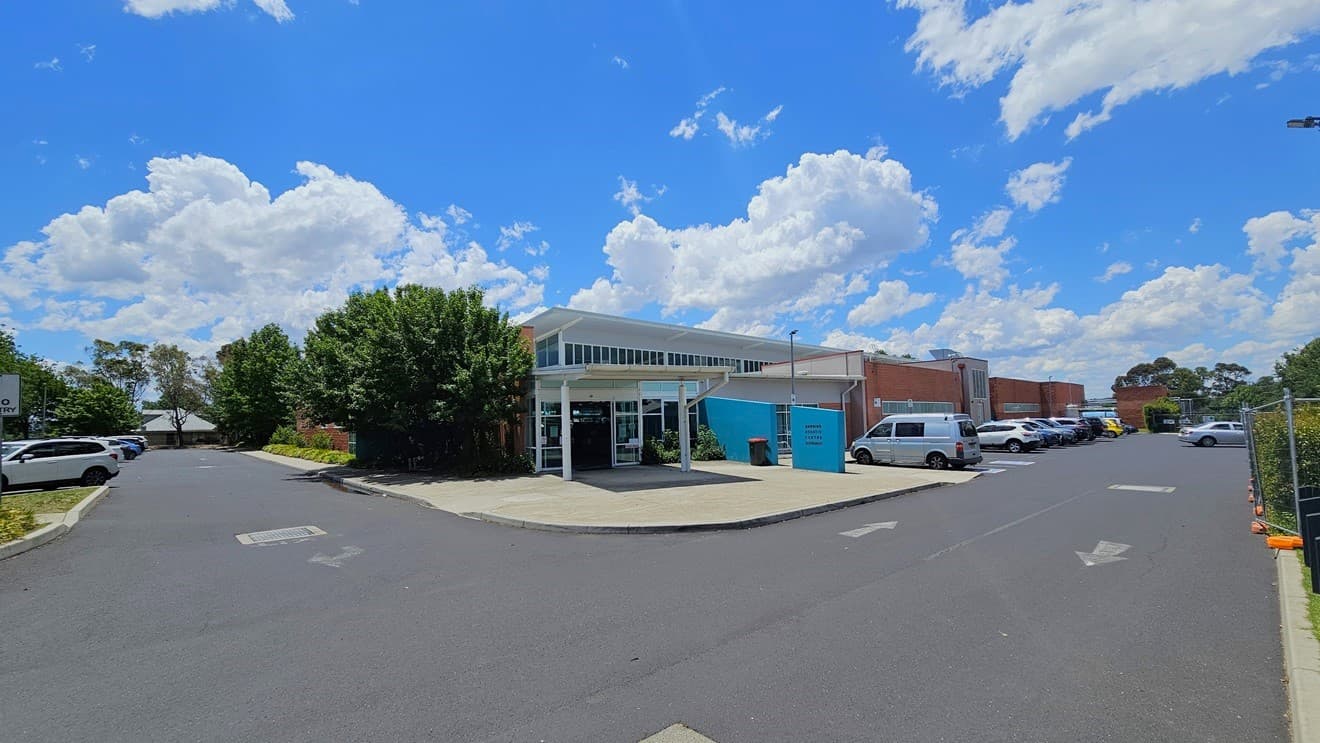 The facility building is a commercial structure featuring red brick, blue-colored concrete, steel, and glass elements. Parking areas, occupied by cars of various colours and models, are situated on both the right and left sides of the building. The surroundings are adorned with trees.