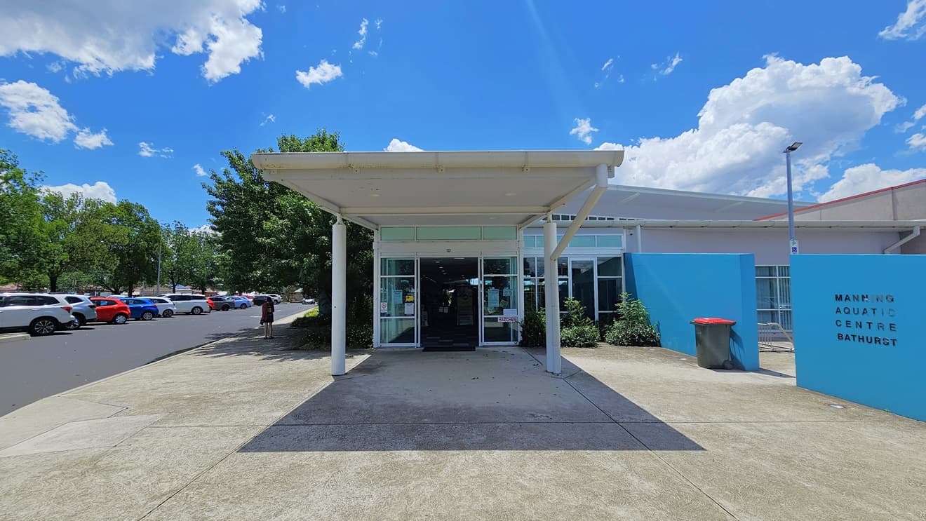 The building entrance features an automatic glass door under a portico. On the left, there is a parking area with cars of various shades and models. To the right is a blue wall displaying the facility's name. A landscape of shrubs is in front of the building, with a rubbish bin and a floodlight positioned on the right near the wall.