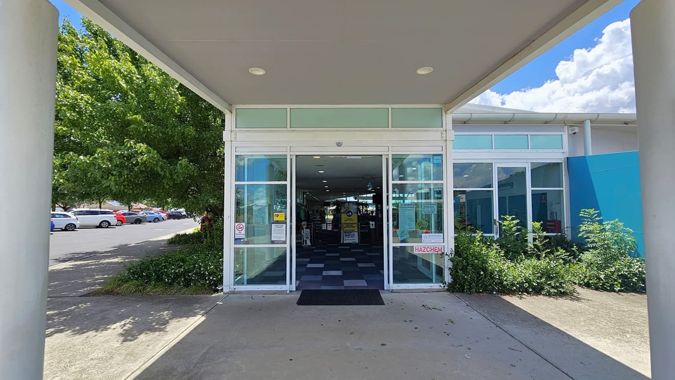 The building entrance has an automatic glass door under a portico, with a non-slip mat on the doorstep. On the left is a parking area with cars of various shades and models. To the right is a blue wall. A landscape of shrubs is in front of the building, with trees and shrubs visible on the left. Inside the building, checked tiles and retail items are visible.