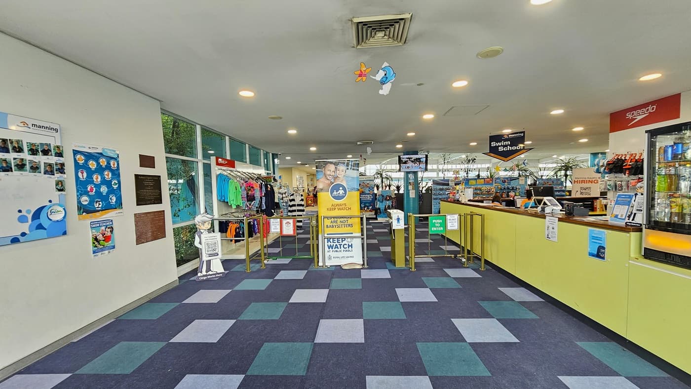 To the right is the reception area with a yellow-front desk, posters, computers, and retail items. A fridge with cold drinks is on its right. A low metal gate serves as a barrier, and a retractable banner stands in the middle. To the left, colorful posters and facility banners adorn the wall. Retail items are displayed after the barrier gates. The ceiling features dolphins and starfish imagery, and the floor has alternating colored carpet tiles.