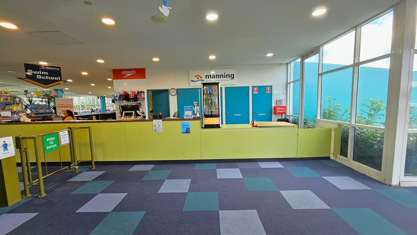 To the right is the reception area with a lower desk, yellow front desk, posters, computers, and floral decorations. Facility banners are also visible. A fridge with cold drinks is behind. A low metal gate serves as a barrier. Retail items are displayed behind the reception desk. The ceiling features dolphins and starfish imagery, and the floor has alternating colored carpet tiles.
