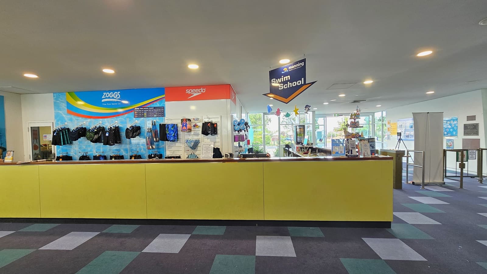 Retail items, including goggles and swimming accessories, are displayed behind the yellow reception desk. To the right is a low gate barrier and a retractable poster. The right wall is adorned with colorful posters and facility banners. The ceiling features all white with LED lights and sea creature imagery, and facility signage is suspended from the ceiling. The floor has alternating colored carpet tiles.