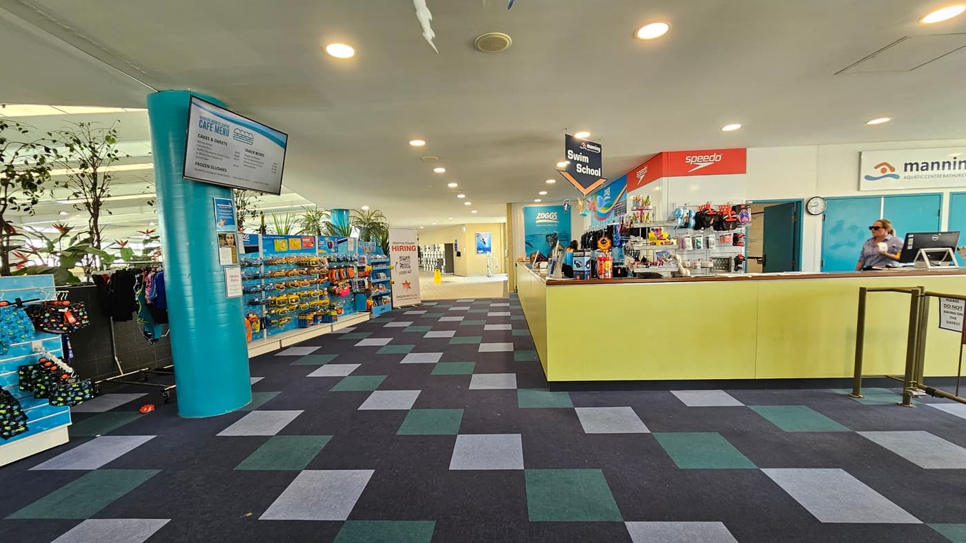 Retail items, such as goggles and swimming accessories, are exhibited behind the yellow reception desk. To the right, there is a low gate barrier and a retractable poster. On the left, tall planter boxes with indoor plants and a long display rack filled with goggles and swimwear are featured. The ceiling is all white with LED lights and facility signage is suspended from the ceiling. A flat-screen TV is mounted to the pillar on the left. The floor is covered with alternating colored carpet tiles