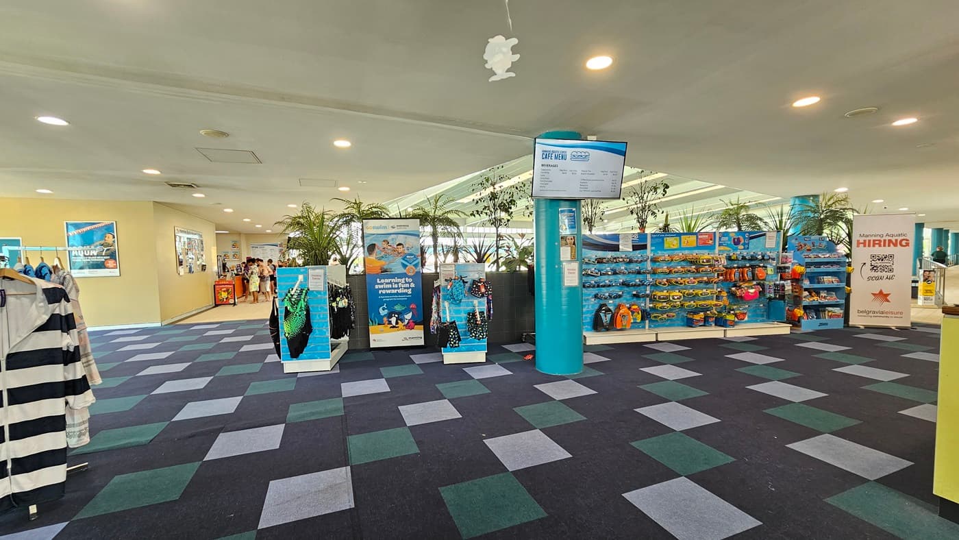 In the middle, tall planter boxes with indoor plants, a display rack filled with goggles, and a swimwear stand. Clothing for sale hangs on the left, and retractable banners are near the planter box. The ceiling is white with LED lights, and facility signage hangs. A flat-screen TV, posters, and people in a corridor on the left are visible. The floor has alternating colored carpet tiles.