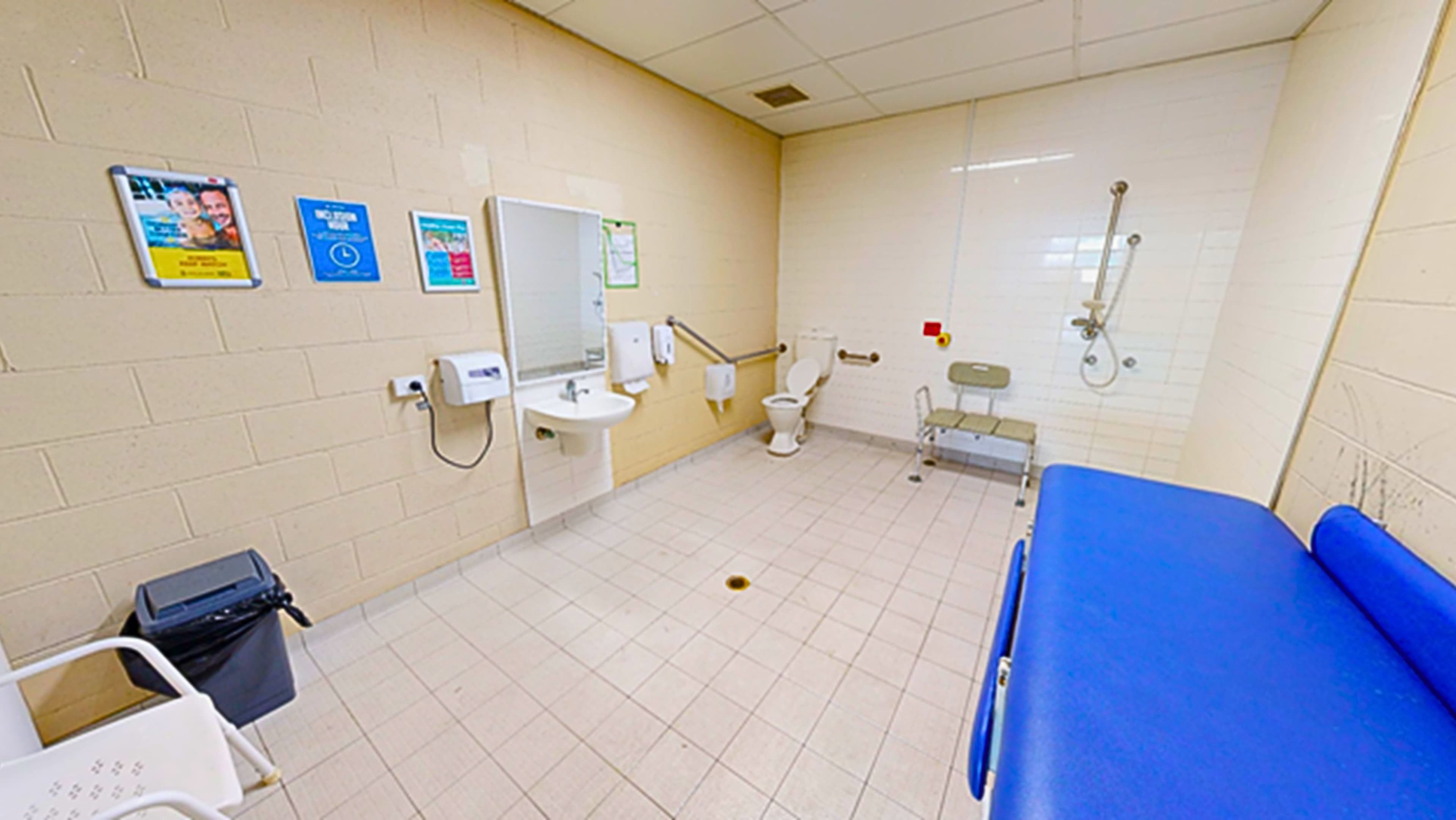 Inside the accessible bathroom, the left corner features a toilet bowl with handrails, a tissue dispenser, and a nearby shower with a bench chair. On the left wall, there's a washbasin, hand dryer, soap and tissue dispenser, and a mirror. Posters are mounted on the wall. A white plastic chair and two rubbish bins are on the left, while a foldable change table is on the right. The floor, ceiling, and wall are covered in white tiles.
