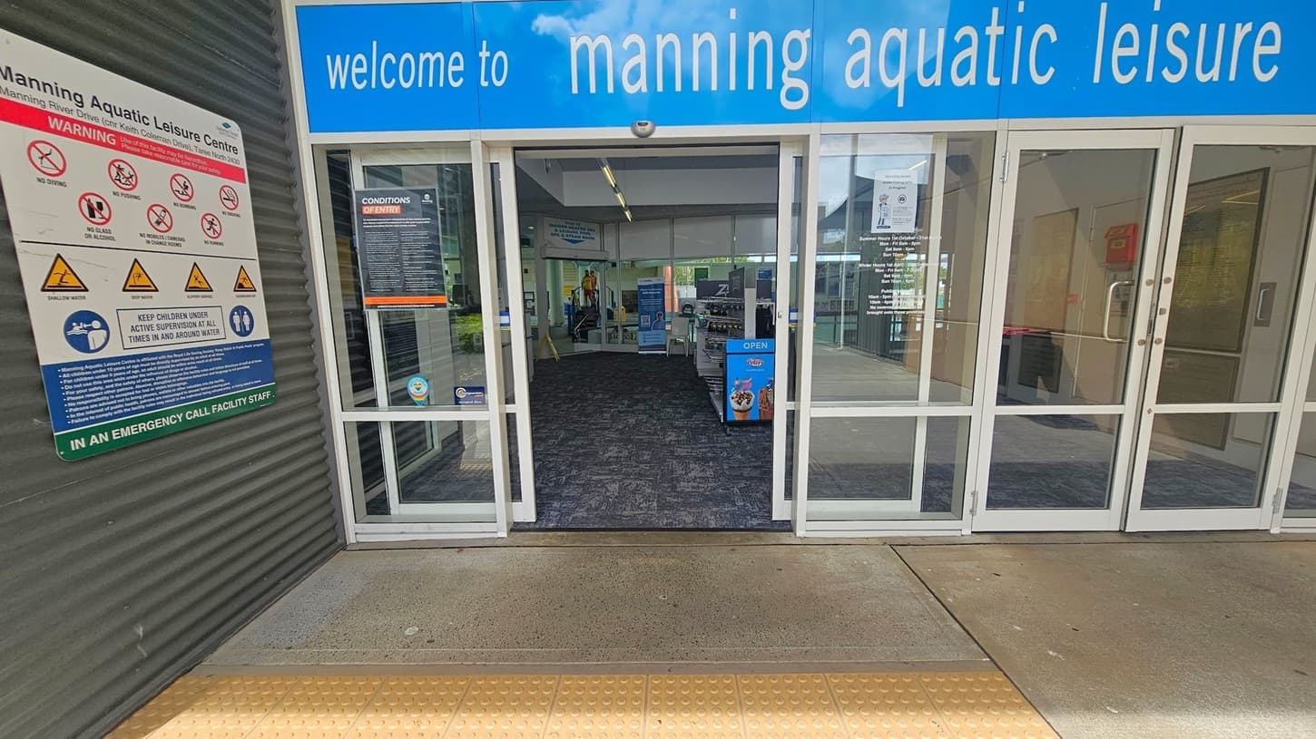 Entrance to Manning Aquatic Leisure Centre with a blue sign above the doors. On the left, a warning sign displays various safety symbols and instructions. The glass doors are open, revealing a carpeted interior with informational signs and a display stand. The floor outside is concrete with a textured yellow strip for accessibility.