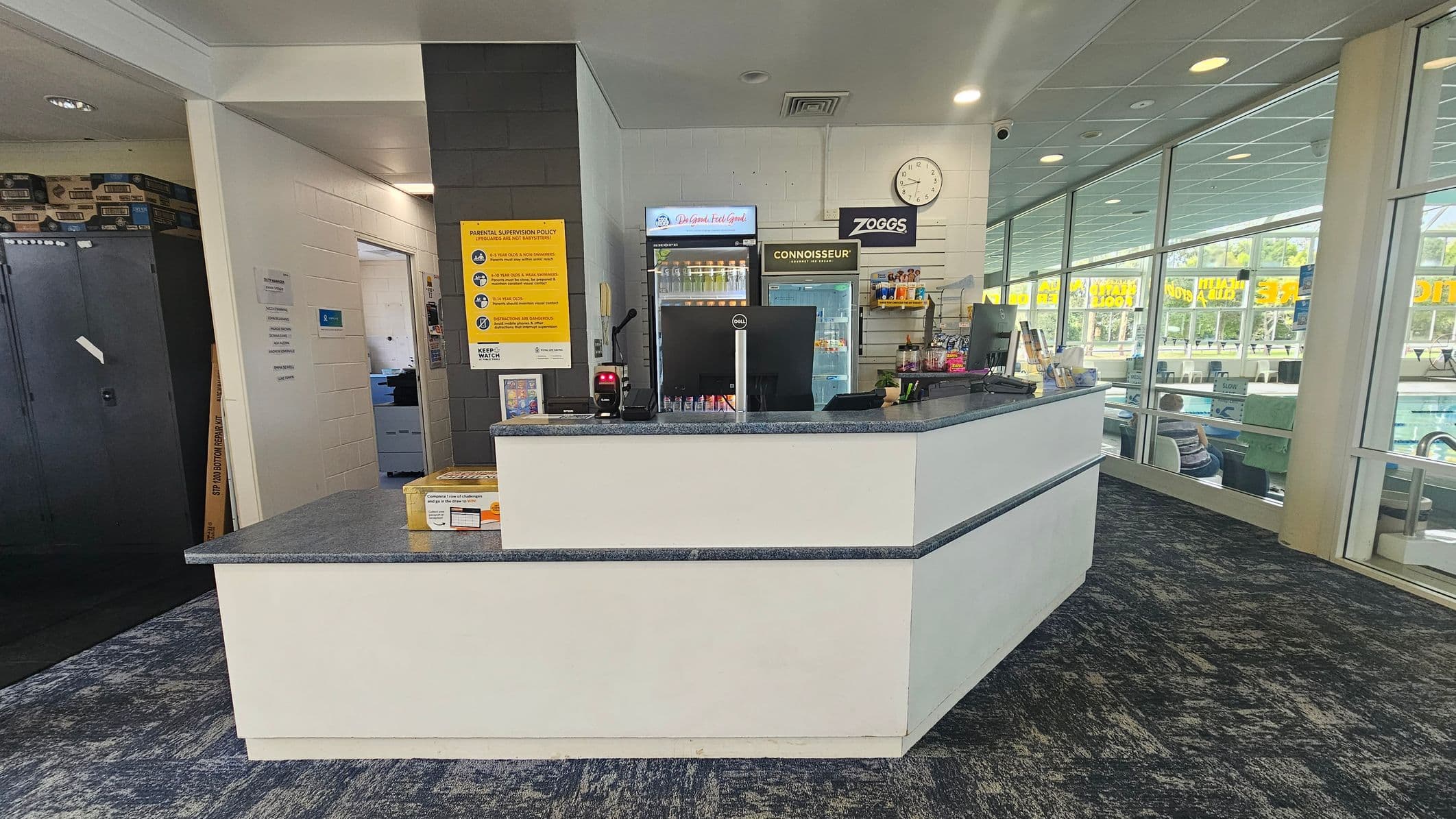 A reception area with a counter in the centre, featuring a computer monitor and a card reader. Behind the counter are two refrigerators stocked with drinks, a clock, and a sign. On the left, there's a doorway leading to another room and a dark cabinet. The right side has large windows showing a pool area with people swimming. The floor is carpeted in a dark pattern, and the ceiling has recessed lighting.