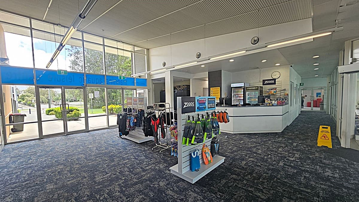 A spacious indoor area with a large glass entrance on the left, allowing natural light to illuminate the space. In the centre, there are display racks with swimming gear and accessories. To the right, a reception counter with a clock above and a drinks fridge behind. The floor is carpeted in a dark pattern, and the ceiling has recessed lighting. A yellow caution sign is visible on the right side near the entrance.