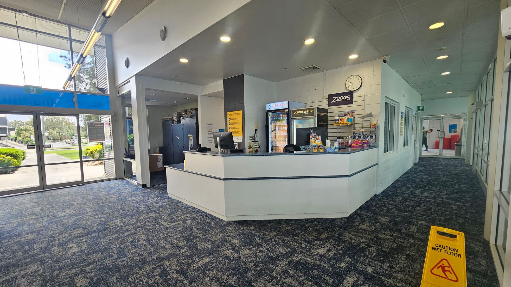 Reception area with a white counter in the centre, displaying snacks and a fridge behind it. A clock and a "Zoggs" sign are on the wall. To the right, a corridor leads to glass doors, with a "Caution Wet Floor" sign on the carpeted floor. On the left, large windows show greenery outside. The ceiling has recessed lighting.