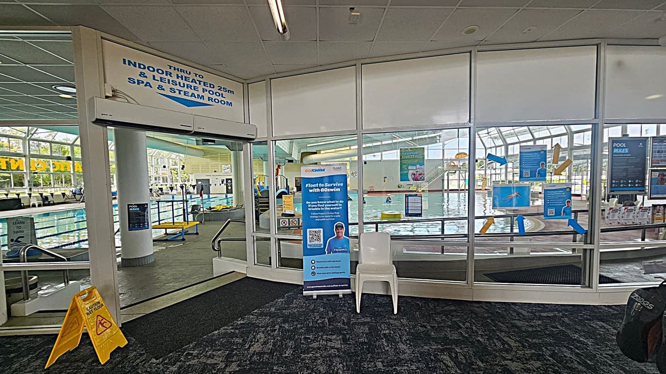 Entrance to an indoor swimming pool area with glass doors and windows. On the left, a sign indicates access to a heated pool, spa, and steam room. A yellow caution sign is on the dark carpeted floor. In the centre, a blue promotional banner and a white plastic chair are visible. Through the glass, the pool area is seen with people swimming and various informational posters on the windows. The ceiling is tiled with fluorescent lighting.