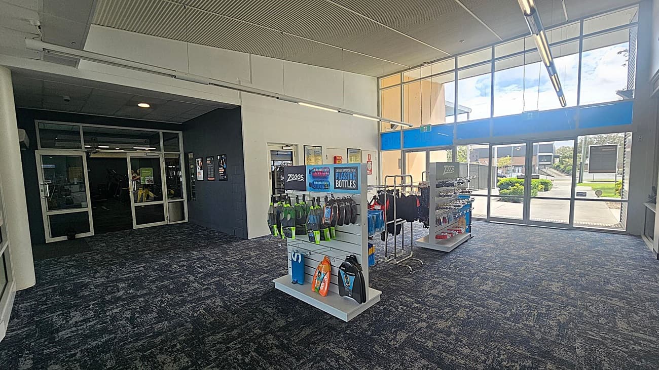 A spacious interior with a dark carpeted floor. On the left, there is a glass door leading to another room, with posters on the adjacent wall. In the centre, a display stand features swimming gear and accessories. To the right, large windows allow natural light to flood in, showing a view of greenery and a building outside. The ceiling has a grid pattern with fluorescent lights.
