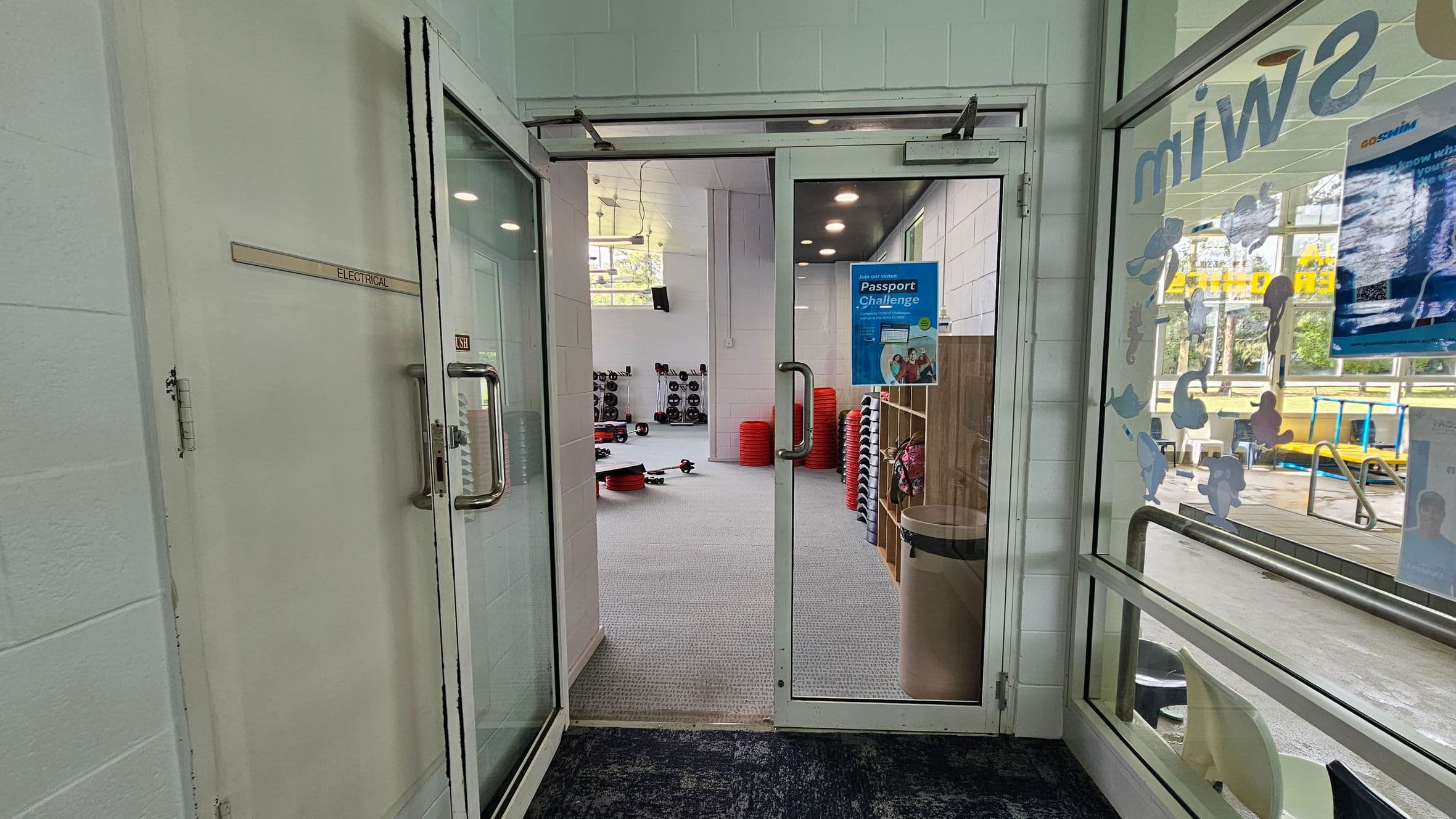 Glass doors open into a gym area with a grey carpeted floor. On the left, there's a door labelled "Electrical." In the middle, stacks of red exercise steps and weights are visible. On the right, a glass wall with aquatic-themed stickers and a view of an outdoor area with chairs and a pool. A poster on the door advertises a "Passport Challenge." The ceiling has recessed lighting.